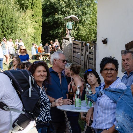 Eine Gruppe von Menschen, darunter Erwachsene und Kinder, versammelt sich im Freien in der Nähe eines Gebäudes. Einige sitzen, andere stehen, und es gibt Flaschen auf einem Tisch. Im Hintergrund sind Bäume und eine Statue zu sehen.