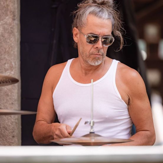 Ein Mann mit grauem Haar und Bart spielt Schlagzeug, trägt ein weißes Tanktop und eine Sonnenbrille.