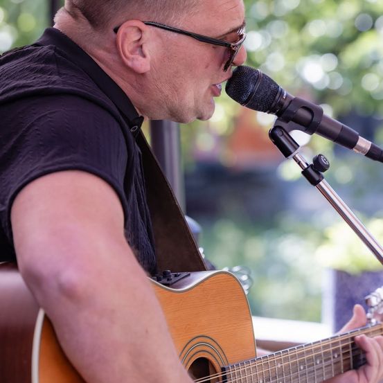 Ein Mann mit Brille und schwarzem Shirt spielt Gitarre und singt in ein Mikrofon.