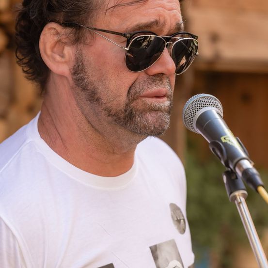 Ein Mann mit Sonnenbrille und weißem T-Shirt spricht in ein Mikrofon, mit verschwommenem Hintergrund.