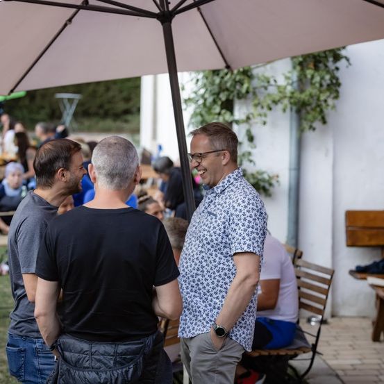 Drei Männer stehen unter einem Schirm bei einer Veranstaltung im Freien, einer lächelt und die anderen schauen zu. Im Hintergrund sind weitere Personen und ein Gebäude zu sehen.