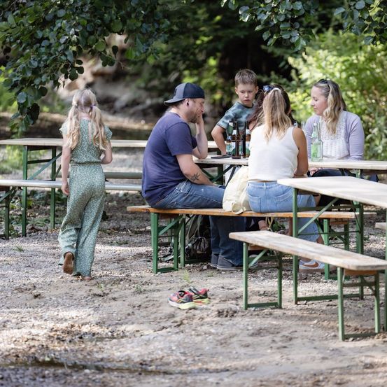 Eine Familie sitzt an einer Picknickbank im Park, eine junges Mädchen steht in der Nähe. Der Mann trägt einen Hut und sieht nachdenklich aus.