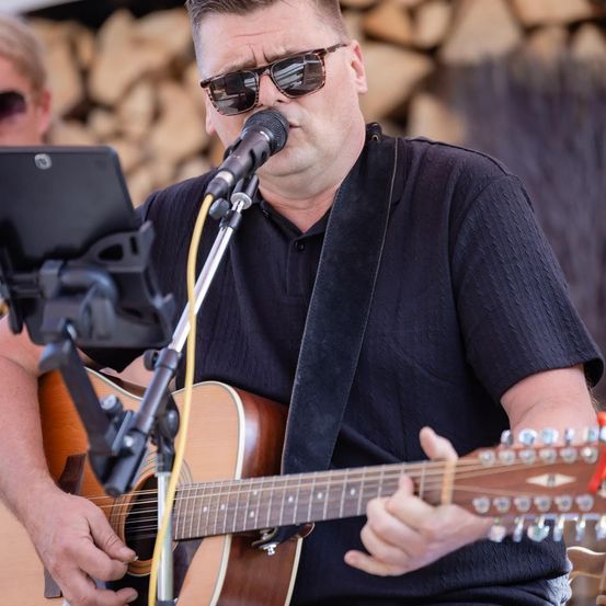 Ein Mann mit Sonnenbrille und schwarzem Shirt spielt Gitarre und singt in ein Mikrofon.