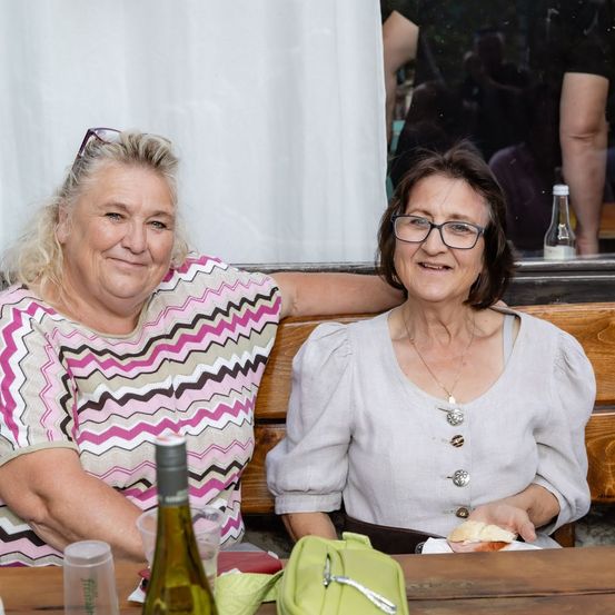 Zwei Frauen sitzen an einem Tisch, eine trägt ein rosa-weiß gestreiftes Shirt und die andere eine graue Bluse, beide lächeln.