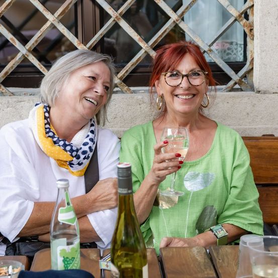 Zwei Frauen sitzen an einem Tisch, eine hält ein Weinglas und lächelt, die andere trägt ein Tuch und schaut sie an. Hinter ihnen ist ein Fenster mit einem Metallrahmen.