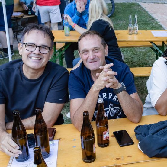 Zwei Männer sitzen an einem Picknicktisch mit Bierflaschen und einem Handy und posieren für ein Foto. Hinter ihnen sitzen andere Leute an Tischen, einige mit Getränken.