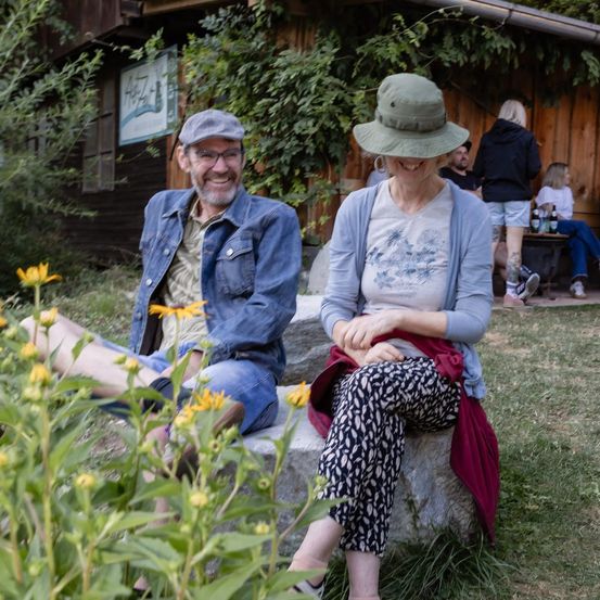 Ein Mann und eine Frau sitzen auf einer Steinbank in einem Garten, lächeln und sind von Blumen umgeben. Hinter ihnen steht ein Holzgebäude mit einem Schild und Leuten, die auf einer Bank sitzen.
