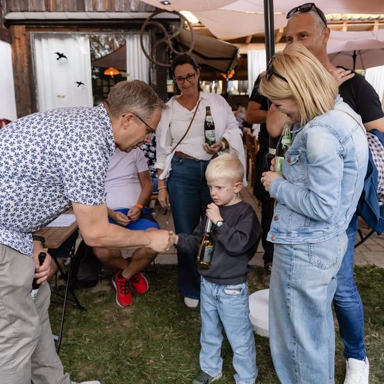 Ein Mann und eine Frau reichen einem kleinen Jungen eine Flasche, während andere im Hintergrund zusehen.