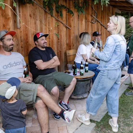 Eine Gruppe von Menschen, einschließlich Erwachsener und Kinder, sitzen auf einer Bank vor einem Holzgebäude. Sie trinken Bier aus Flaschen und Gläsern. Eine Frau steht und hält eine Flasche Bier.