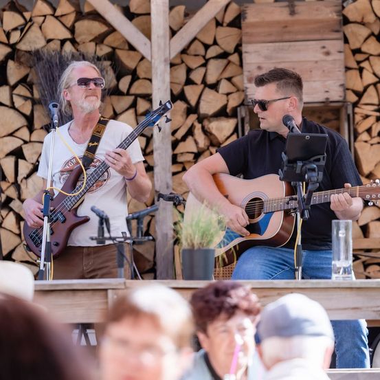 Zwei Männer spielen Gitarren auf einer Bühne vor einem Haufen Holzscheite, während eine Menge zuschaut.