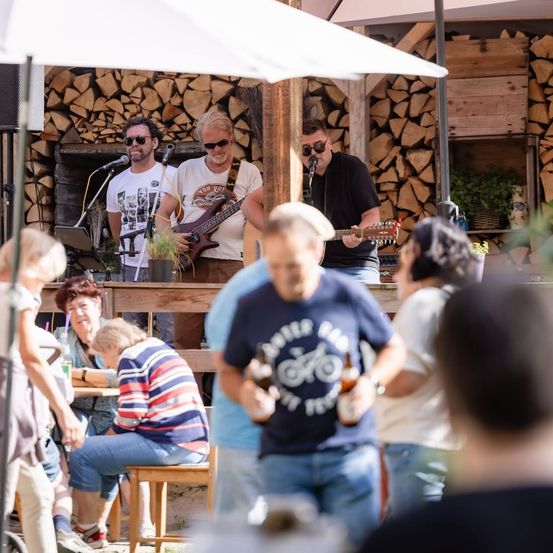 Eine Gruppe Musiker tritt auf einer Bühne in einer Außenumgebung auf. Sie spielen Gitarren und singen in Mikrofone. Das Publikum sitzt an Tischen und genießt die Musik.