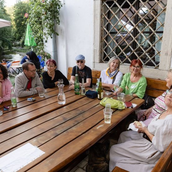 Eine Gruppe von Menschen sitzt um einen Holztisch in einer Außenumgebung, einige mit Brille und lächelnd. Flaschen, Gläser und andere Gegenstände sind auf dem Tisch. Hinter ihnen befindet sich ein Fenster mit einem Metallgitter und einem grünen Schirm.