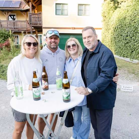 Vier Personen stehen um einen Tisch mit Freibier-Flaschen und posieren lächelnd für ein Foto. Hinter ihnen steht ein Haus mit Solarpanelen auf dem Dach.