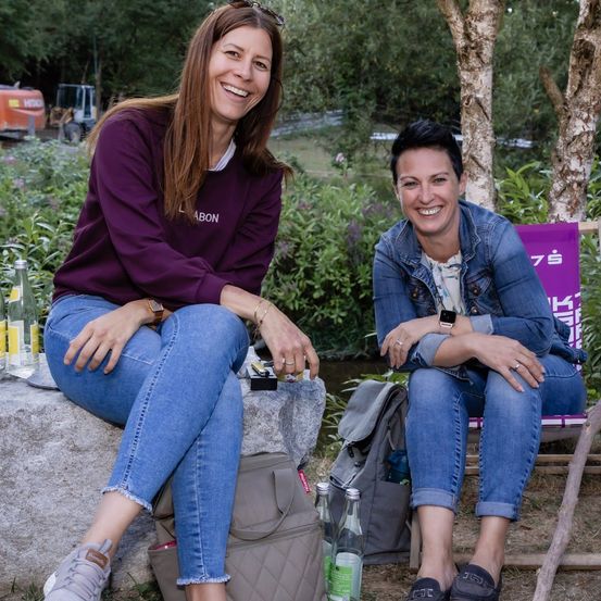 Zwei Frauen sitzen auf einem Felsen und einem Stuhl, lächeln und posieren für ein Foto. Sie tragen Freizeitkleidung und haben Taschen und Flaschen um sich herum. Im Hintergrund sind Bäume und Pflanzen verschwommen zu sehen.