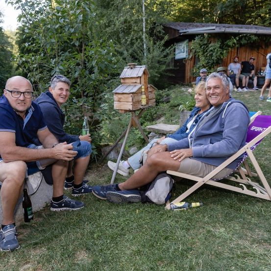 Eine Gruppe von Menschen sitzt auf Liegestühlen in einem Garten, lächelt und posiert für ein Foto. Ein hölzerne Vogelhaus steht in der Nähe.