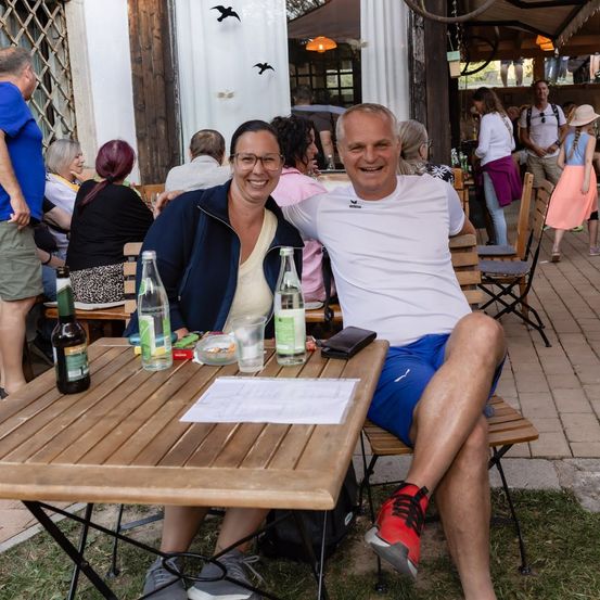 Ein Mann und eine Frau sitzen auf einer Bank vor einem Restaurant, lächeln und posieren für ein Foto. Auf dem Tisch vor ihnen befinden sich Flaschen, eine Geldbörse und ein Blatt Papier.