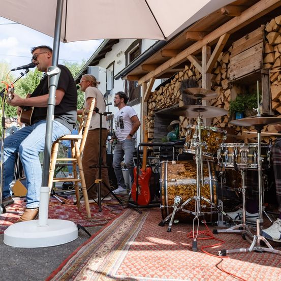 Eine Band spielt auf einer Bühne mit einem Holzhaus im Hintergrund, bestehend aus einem Gitarristen, einem Schlagzeuger und einem Sänger unter einem Regenschirm.