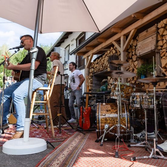 Eine Band spielt im Freien, mit einem Gitarristen, der unter einem Regenschirm sitzt, einem Schlagzeuger mit Becken und einem Saxophonisten, der dahinter steht. Die Umgebung umfasst eine Holzstruktur mit gestapelten Holzscheiten und ein Holzhaus.