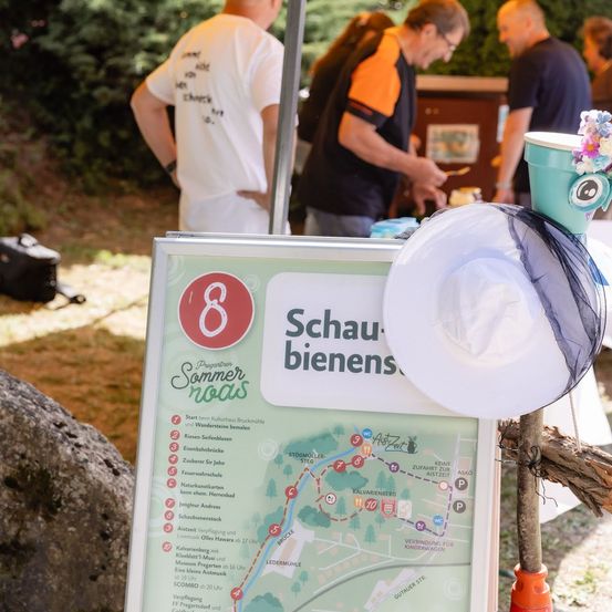 Eine Karte mit einem Schild, das 'Schaubienens' und die Nummer '8' zeigt, ist auf einem Ständer ausgestellt. Menschen stehen hinter der Karte, einer von ihnen trägt eine Uhr. Ein Hut mit einer Blume darauf ist auf dem Ständer platziert.