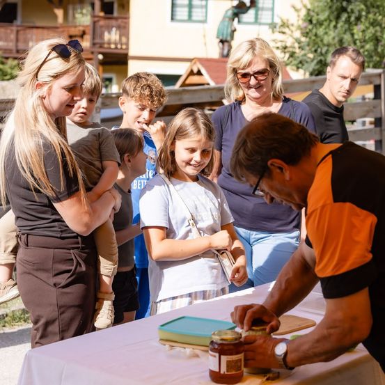 Eine Familie versammelt sich um einen Tisch, an dem ein Mann eine Glas mit Marmelade vorbereitet. Kinder und Erwachsene beobachten ihn. Der Ort ist im Freien mit einem Holzzaun und einem Haus im Hintergrund.