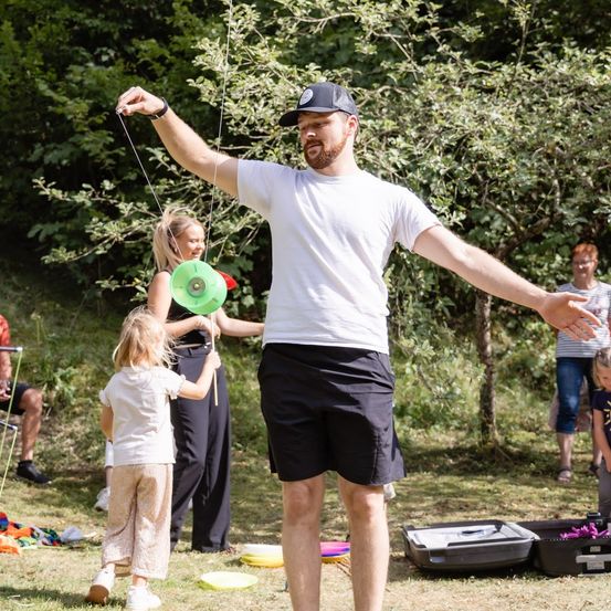 Ein Mann in einem weißen T-Shirt und schwarzen Shorts fliegt einen grünen Frisbee, während ein junges Mädchen am Seil zieht. Eine Frau steht hinter ihnen, und andere Menschen sind im Hintergrund.