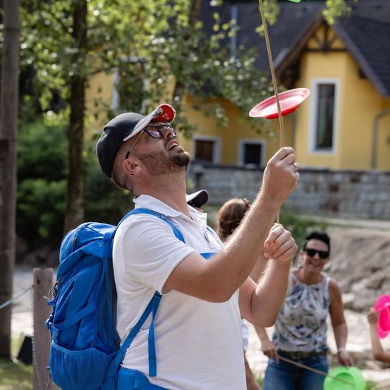 Ein Mann mit blauem Rucksack und Mütze jongliert mit einem roten Frisbee im Freien. Hinter ihm steht eine Frau in einem ärmellosen Shirt, die lächelt und einen pinkfarbenen Frisbee hält. Im Hintergrund befinden sich ein gelbes Haus und Bäume.