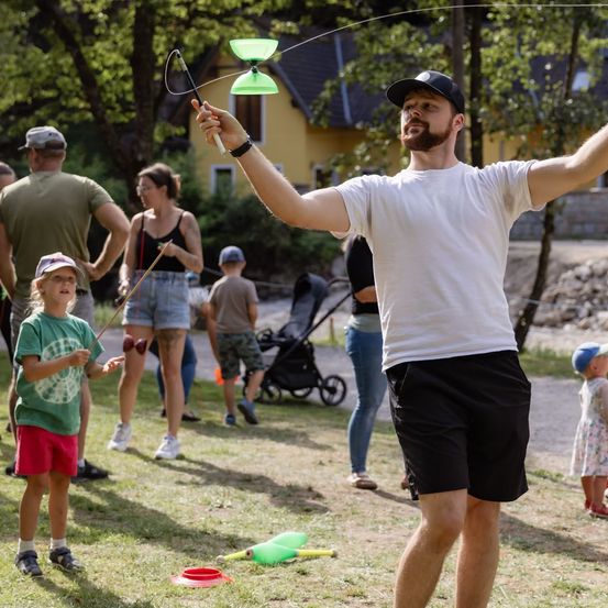 Ein Mann in einem weißen T-Shirt und schwarzen Shorts spielt mit einem grünen Drehteller, während ein junges Mädchen ein ähnliches Spielzeug in einem Grasbereich mit anderen Leuten und einem Haus im Hintergrund hält.