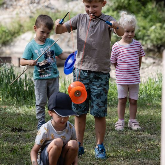 Drei Kinder spielen mit bunten Frisbees in einem Grasbereich, ein Junge hält einen Stock und zwei Frisbees, ein anderer Junge kniet mit einer Mütze, und ein Mädchen steht hinter ihnen.