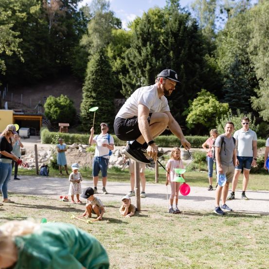 Ein Mann springt über einen Pfosten und hält dabei einen Frisbee, umgeben von spielenden Menschen und Kindern in einem Park.