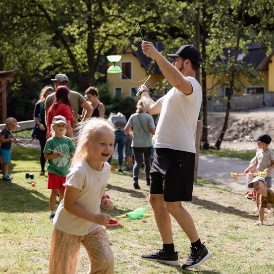 Ein Mann und ein junges Mädchen spielen mit grünen Spielzeug-Drechseln in einem Park, umgeben von anderen Kindern und Erwachsenen.
