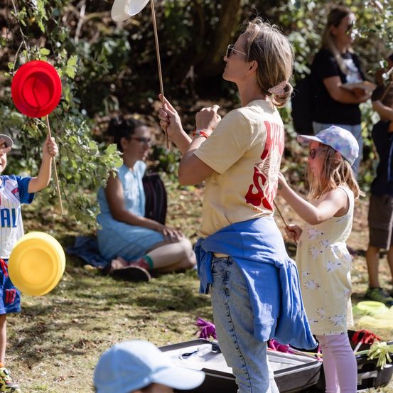 Eine Frau und zwei Kinder spielen mit roten und gelben Frisbees im Park. Die Frau hält einen Stock mit einem weißen Frisbee. Ein Mädchen hält ein rotes Frisbee. Ein Junge hält ein gelbes Frisbee.