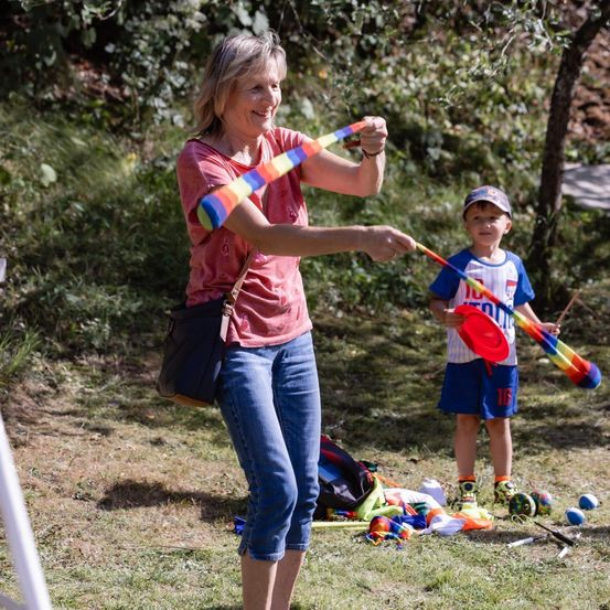 Eine ältere Frau und ein junger Junge spielen mit bunten Bändern in einem äußeren Grasbereich. Die Frau lächelt und hält ein Band, während der Junge einen roten Frisbee hält.