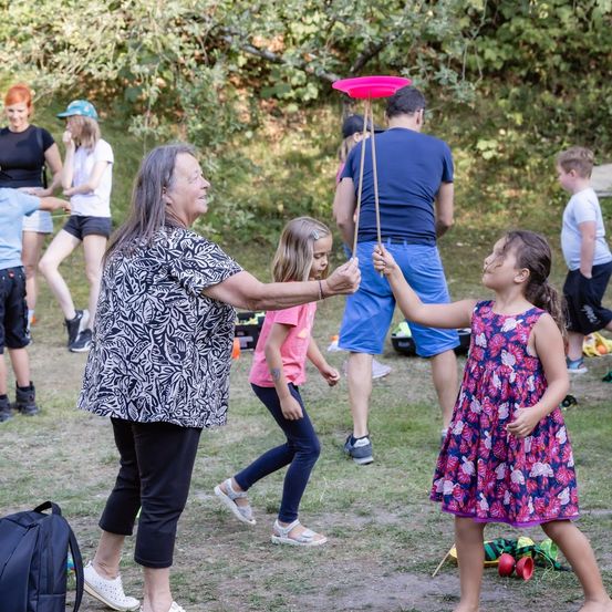 Eine ältere Frau und ein junges Mädchen spielen mit einem rosa Frisbee auf einem Rasenbereich, während andere Kinder und Erwachsene im Hintergrund sind.