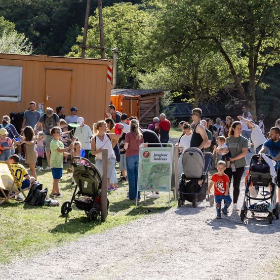 Eine Gruppe von Menschen, einschließlich Familien mit kleinen Kindern, versammelt sich in einem Park mit einem Kartenschild und einem Gebäude im Hintergrund.
