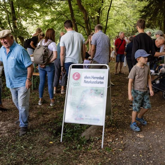 Eine Gruppe von Menschen geht in einem Wald, einige tragen Rucksäcke und andere schieben Kinderwagen. Ein Schild mit einer Karte ist auf dem Boden aufgestellt.