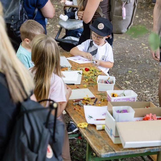 Eine Gruppe von Kindern ist um einen Tisch versammelt und beschäftigt sich mit einer Kunstaktivität. Ein Kind trägt Hut und Brille. Verschiedene Kunstmaterialien sind auf dem Tisch verstreut.