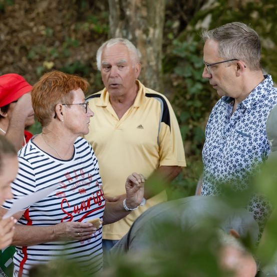 Eine Gruppe älterer Erwachsener versammelt sich in einem Wald, wobei eine Frau ein gestreiftes Shirt trägt und ein Mann ein gelbes Shirt.