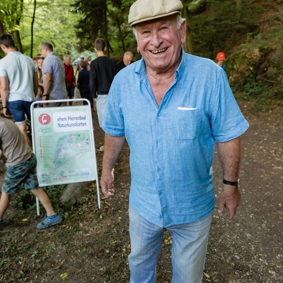 Ein älterer Mann in einem blauen Hemd und Hut lächelt in Richtung Kamera in einer Waldumgebung, mit einem Kartenschild hinter ihm und mehreren Personen im Hintergrund.