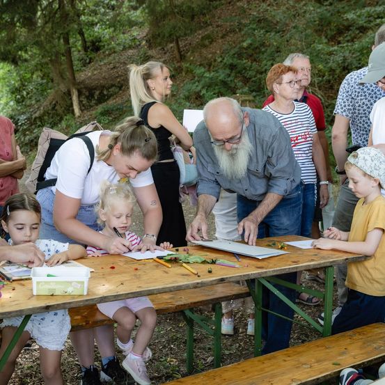 Eine Gruppe von Menschen, darunter Erwachsene und Kinder, versammelt sich um einen Tisch in einer Waldumgebung. Ein Mann mit langem Bart hilft einem kleinen Mädchen bei ihrem Zeichnen, während eine andere Frau einem Kleinkind hilft. Verschiedene Zeichenutensilien liegen auf dem Tisch.