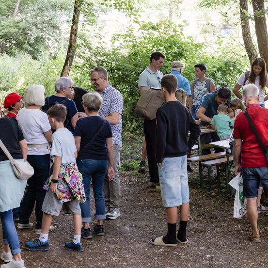 Eine Gruppe von Menschen, einschließlich Erwachsener und Kindern, versammelt sich in einem bewaldeten Bereich, einige stehend und andere sitzend an einem Picknicktisch. Eine Person hält eine weiße Tasche.