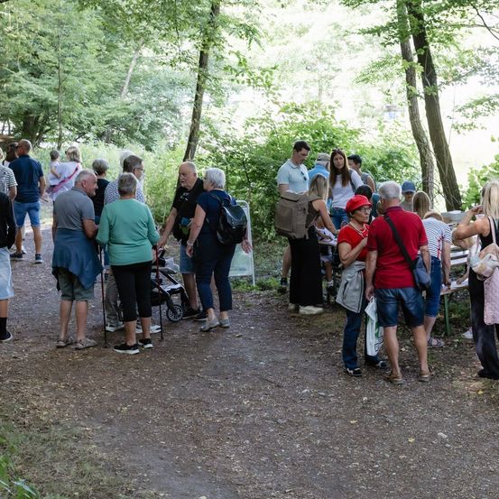Eine Gruppe von Menschen, einige mit Rucksäcken, versammelt sich in einem bewaldeten Gebiet, möglicherweise in Erwartung von etwas. Einige stehen, andere sitzen auf einer Bank.