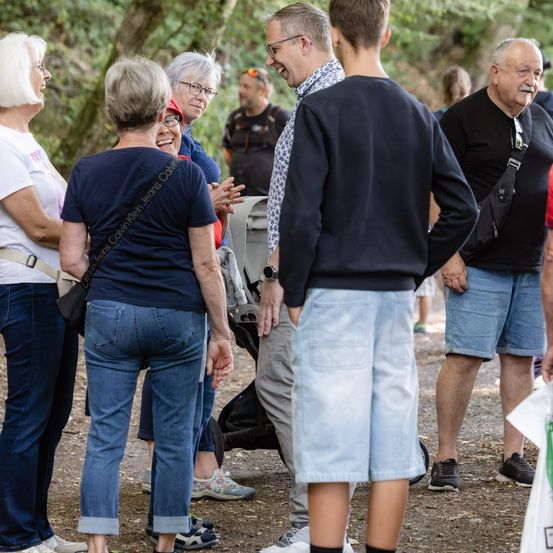 Eine Gruppe von Menschen, darunter eine Frau in einem blauen Shirt und ein Mann mit einem roten Hut, stehen und unterhalten sich in einem Wald.