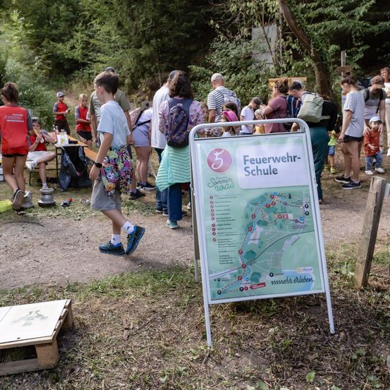 Eine Gruppe von Menschen versammelt sich in einem Waldgebiet, einige gehen, andere sitzen auf Bänken. Ein Kartenschild steht auf dem Boden, und eine Person geht darauf zu.