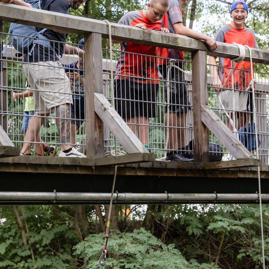 Drei junge Männer in orangefarbenen und grauen T-Shirts stehen auf einer Holzbrücke mit Metallgeländern, einer von ihnen lehnt sich an das Geländer.