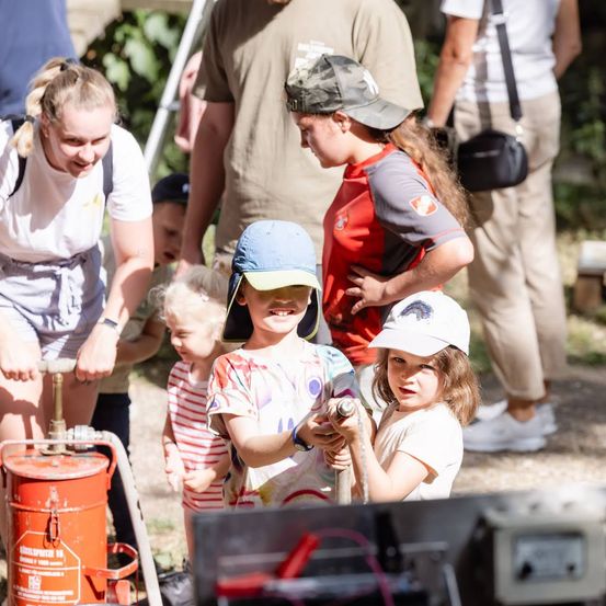 Eine Gruppe von Kindern und Erwachsenen ist draußen versammelt, einige halten Werkzeuge und lächeln. Ein Kind hält einen Schlauch und scheint etwas zu gießen.