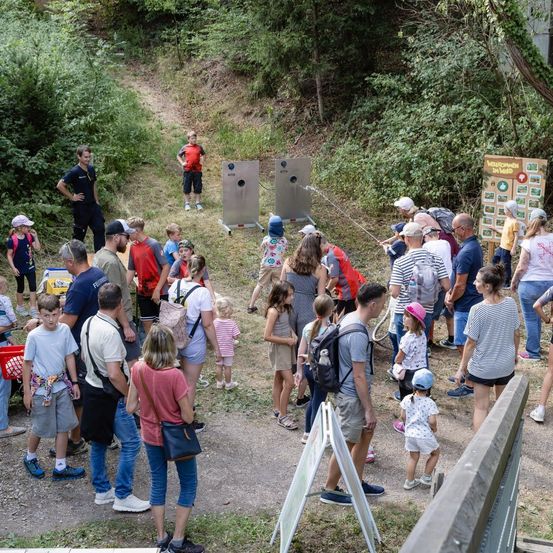 Eine Gruppe von Menschen, darunter Erwachsene und Kinder, versammelt sich in einer parkähnlichen Umgebung. Einige beobachten eine Zielschießaktivität, während andere in Gespräche vertieft sind. Das Gebiet ist von viel Grün umgeben und hat einige Schilder.