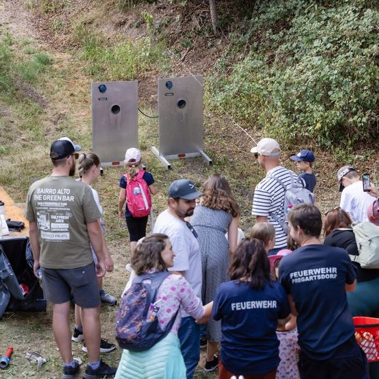 Eine Gruppe von Menschen steht auf einem Feld, einige tragen Hüte und Turnschuhe. Sie schauen auf zwei Metallziele mit Löchern. Einige von ihnen halten einen Rucksack. Es gibt einen Tisch mit einer Flasche und anderen Gegenständen darauf.