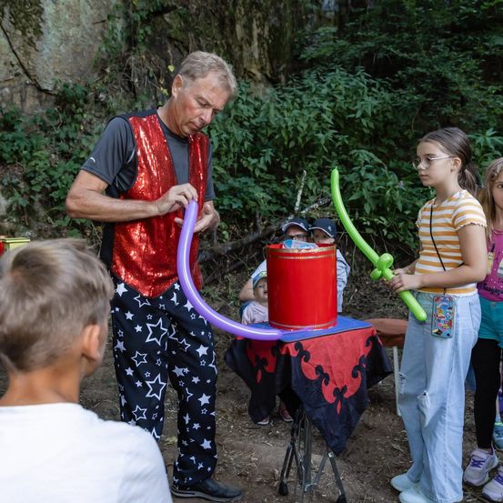 Ein Mann in einer roten Weste und Sternenhosen führt einen Ballontrick mit einem roten Hut vor Kindern durch.