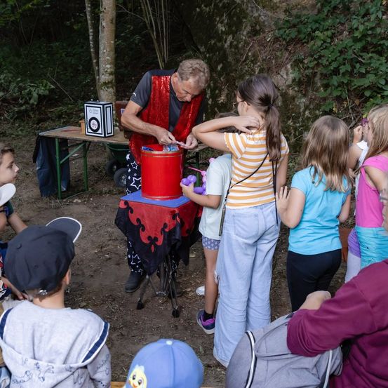 Ein Zauberer führt einen Trick mit einem roten Eimer vor einer Gruppe von Kindern in einem Wald vor.