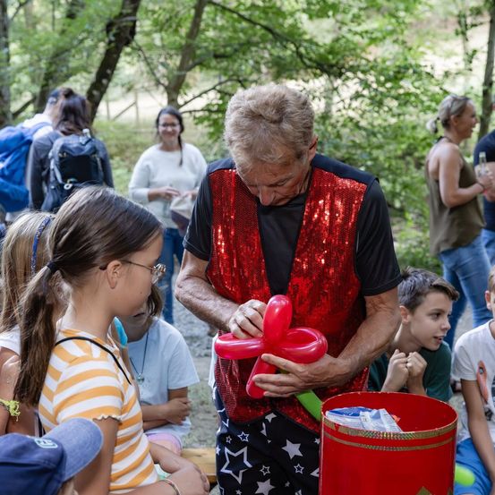 Ein Mann in einem roten Paillettenweste macht eine Ballonfigur für ein junges Mädchen in einem gestreiften Shirt.